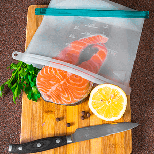 A salmon steak, lemon, and herbs halfway inside of a reusable food bag. All of these items are on top of a chopping board with a sharp knife,
