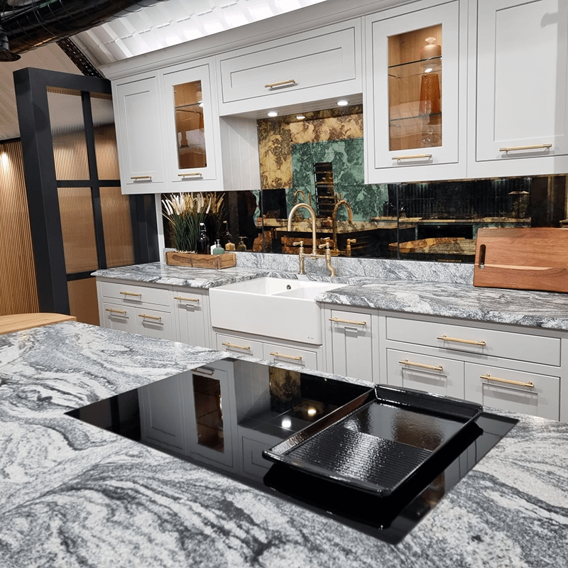 A black induction hob built into a grey marble island counter, with a white and grey toned kitchen in the background with gold hardware.