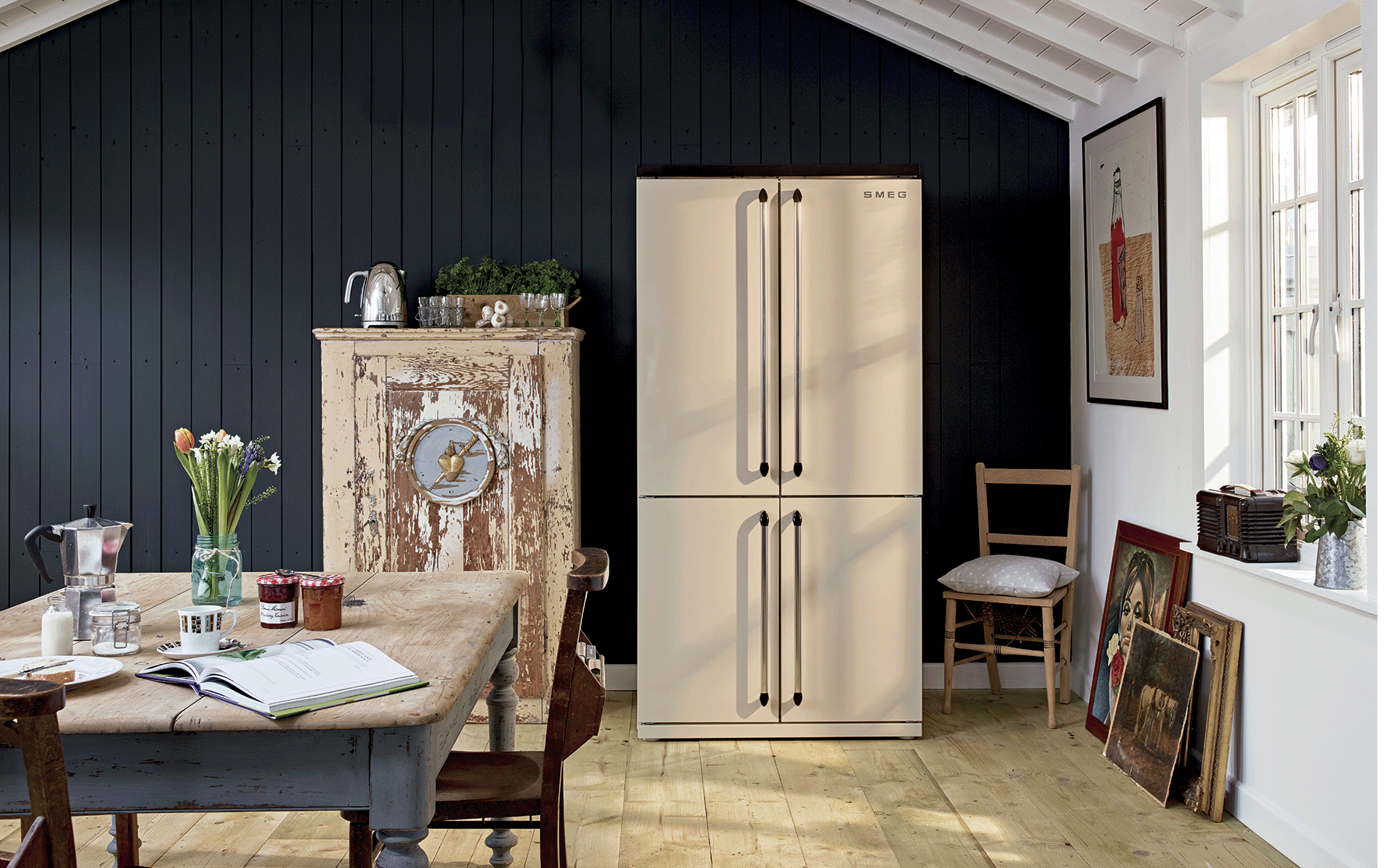 Cream Smeg American Fridge Freezer in a room with a blue distressed dining table and paintings on the floor.