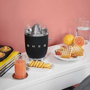 Black Smeg citrus juicer next to various food items and peach coloured juice in a glass.