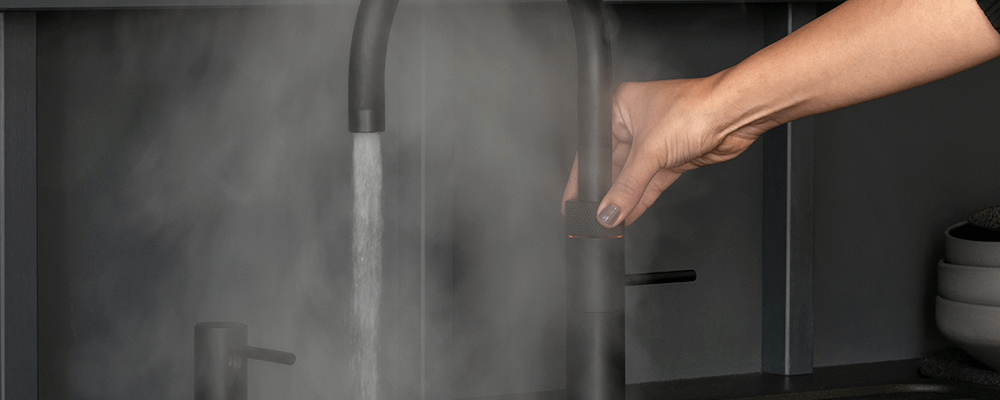 Hand shown turning on a round black boiling water tap with steam all around.