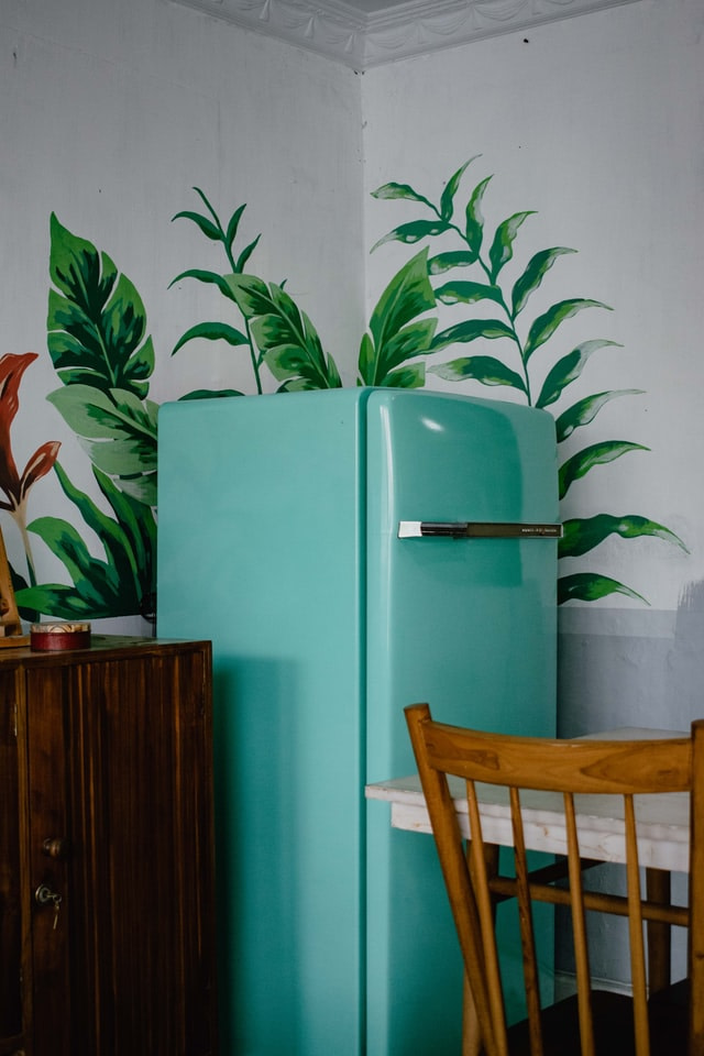 teal fridge by a table and chair against a white wall with plant decal