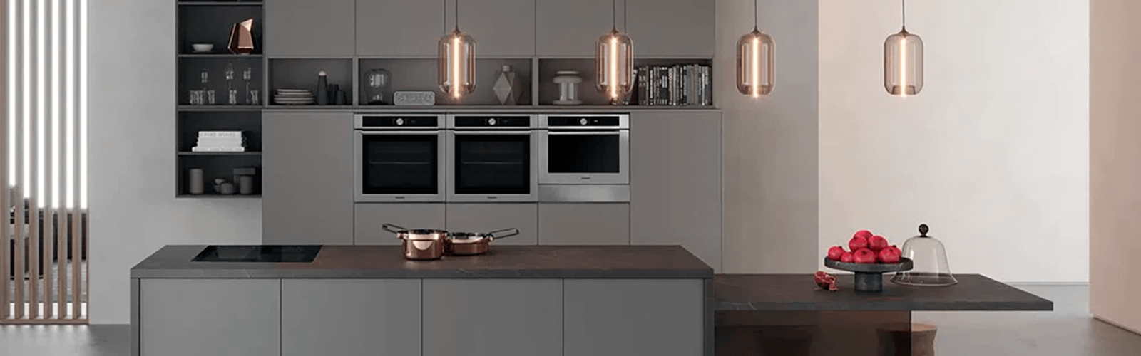 Grey kitchen with 3 built in ovens, a Hotpoint induction hob, hanging pendant lights and rose gold pans on the counter.