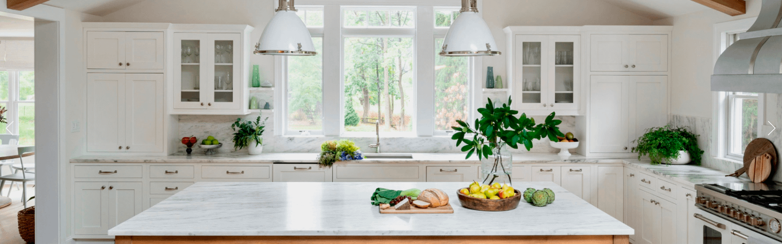 Large white kitchen with white pendant lights and lots of plants. Fresh ingredients are on top of a white marble island. A Bertazonni range cooker is on the right.