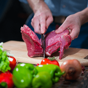 Cutting meat on a wooden chopping board, surrounded in red and green peppers