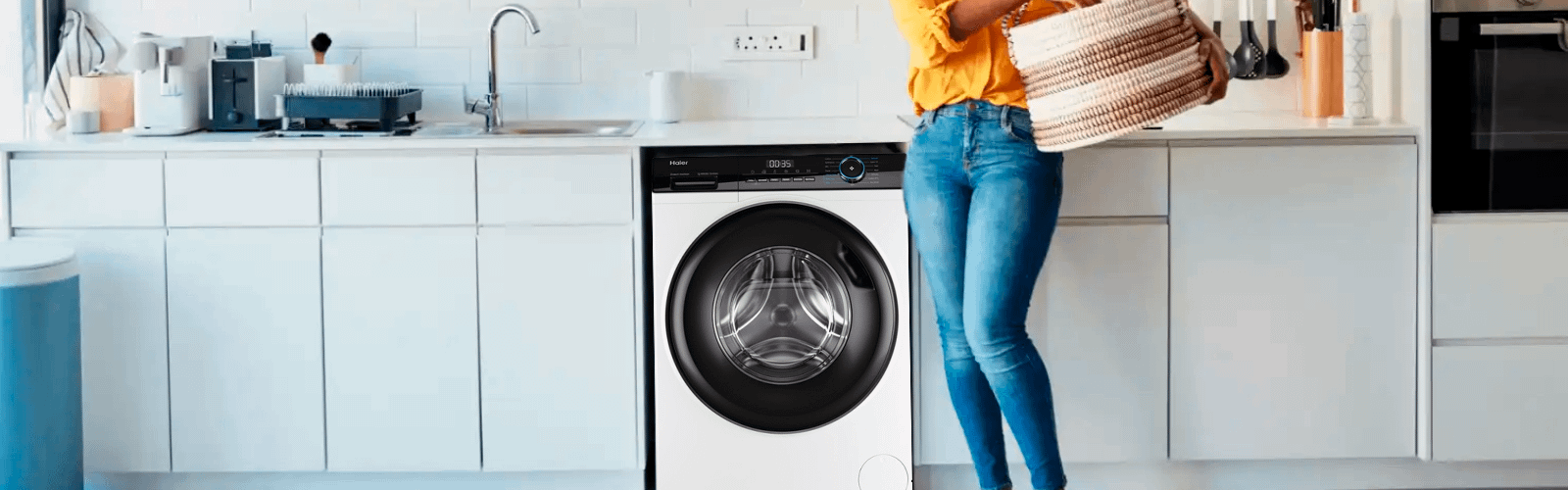Haier washing machine in a white kitchen. Woman in jean and yellow top is holding a basket in front.
