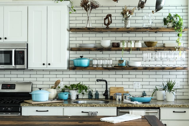 pale green brick wall with wooden shelves that hold pans and kitchen utensils above a kitchen coutertop