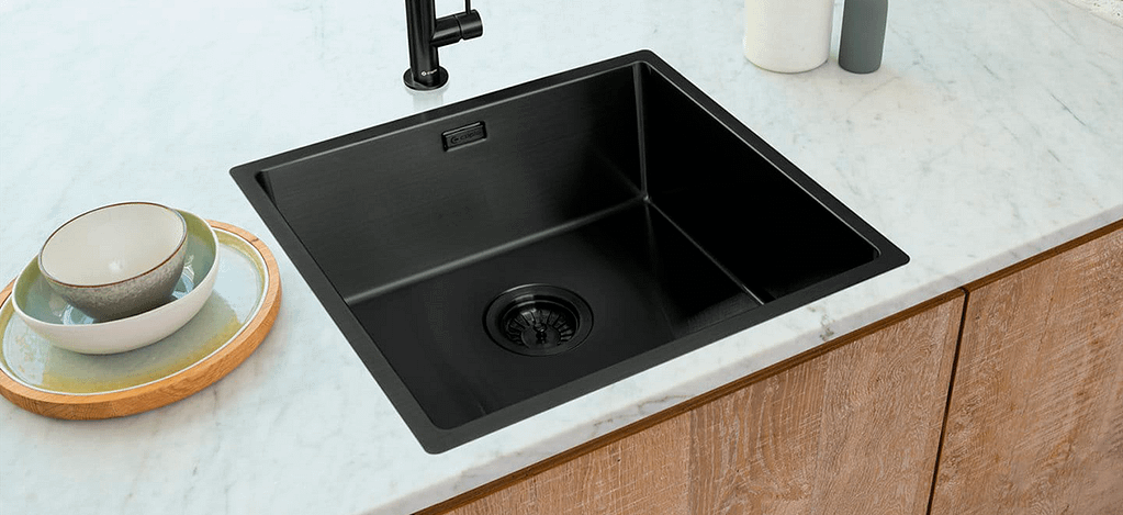 A dark gunmetal stainless steel sink installed in a white marble countertop, with some clean plates piled next to it.