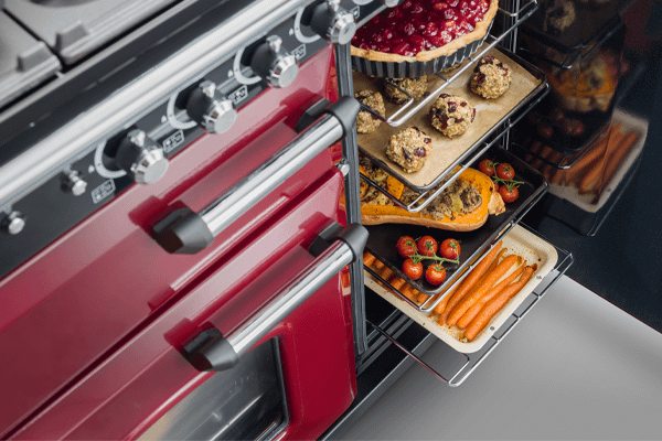A zoomed in cranberry Rangemaster range cooker with the right door ajar and displaying a range of food, including a cherry tart, cookies, butternut squash and carrots