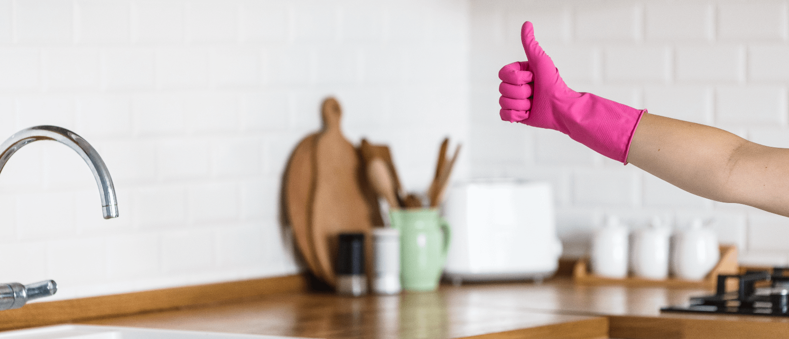 Hand giving the thumbs up whilst wearing a hot pink kitchen glove. Background shows wooden kitchen countertop with stainless steel tap and sink.
