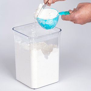 Container full of flour, with person using a blue measuring cup for a cup of flour.