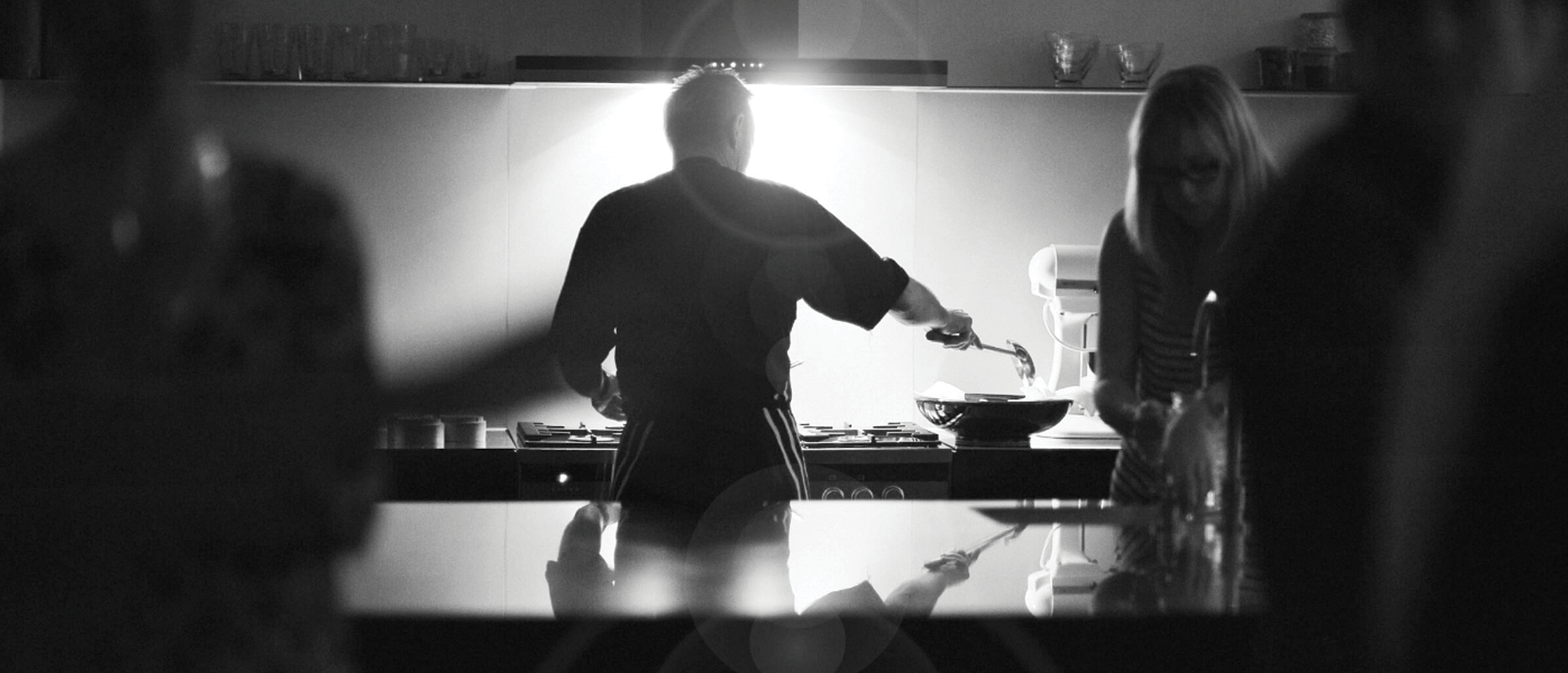 Black and white image of a busy kitchen. Man is seen standing at a range cooker scooping food into a bowl. Woman filling jug with water from sink.