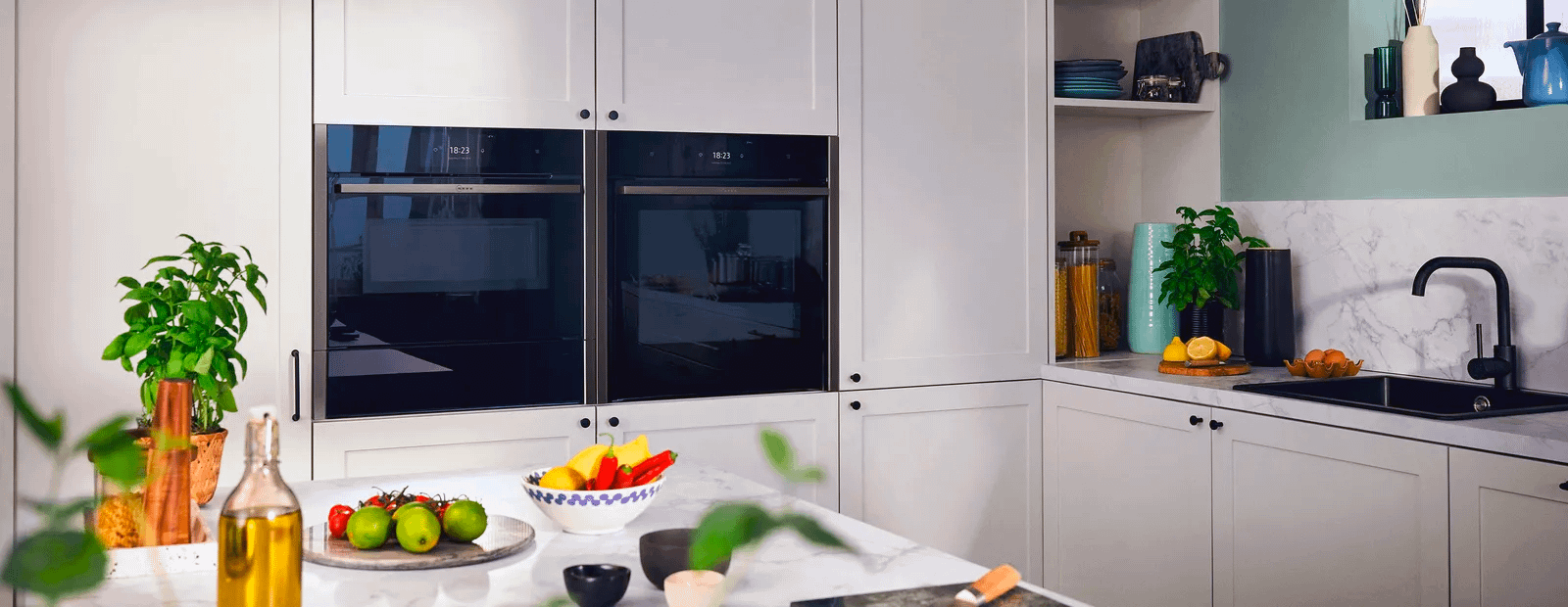 White kitchen with two built in Neff ovens, bright fruit and veg on the counters, and a black sink.