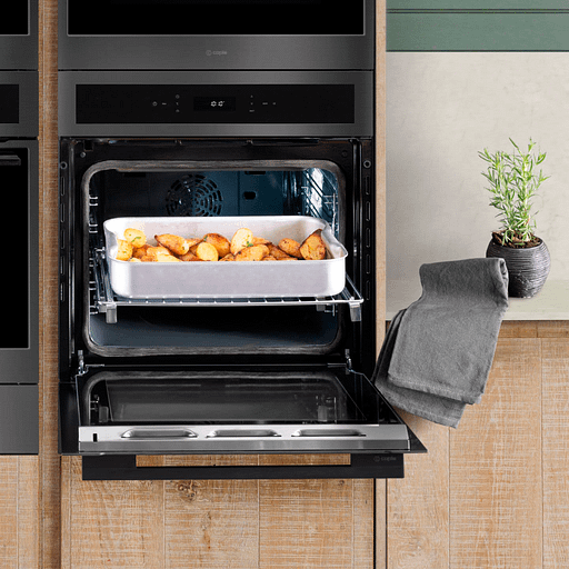 A gunmetal finished oven built into wooden cabinetry, with open oven cavity to reveal a dish of roast potatoes.