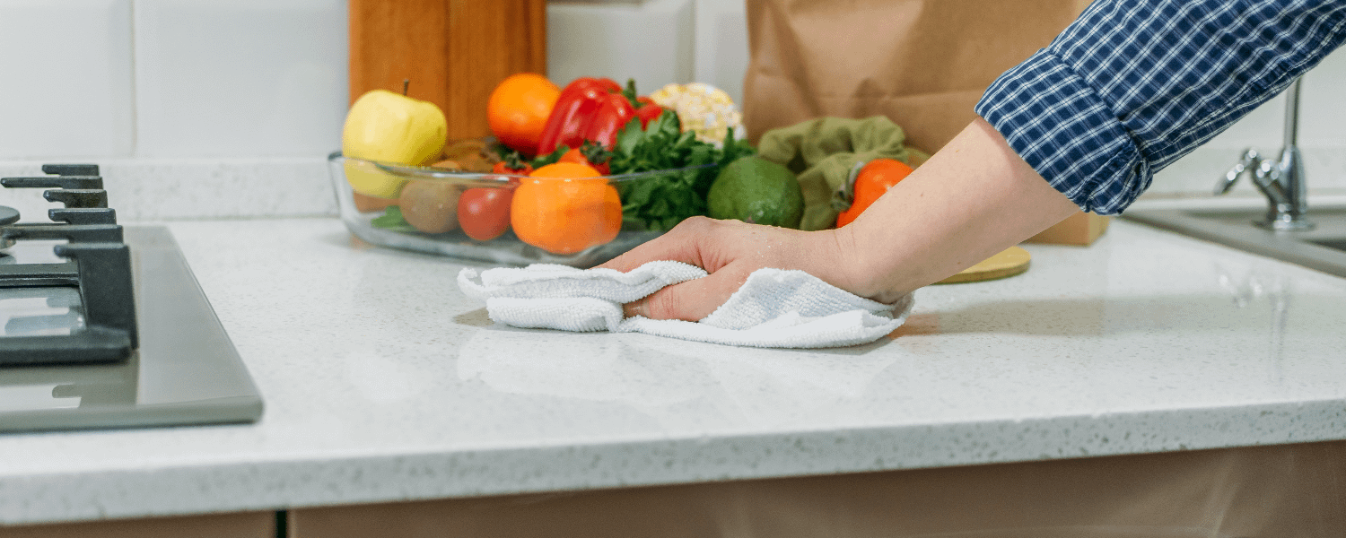 Hand wiping kitchen counter with a white clothes. Colourful fruit and vegetables are in a bowl in the background.