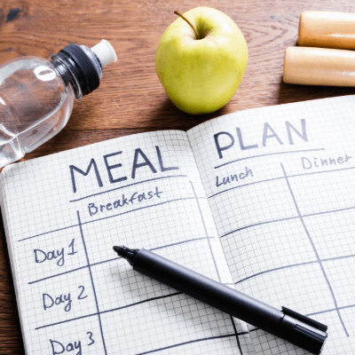 Notebook with a meal plan table drawn. A waterbottle and apple are next to the notebook. A black pen is on top.