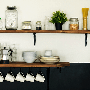 Wooden kitchen shelves on a white wall. Filled with plates, glasses, and jars of food items. White mugs hang from the bottom shelf.