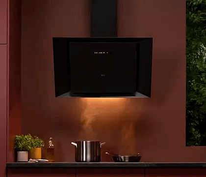 AEG cooker hood above steaming pot and pan. Dark room shows the lights from the hood glowing down against the orange wall behind