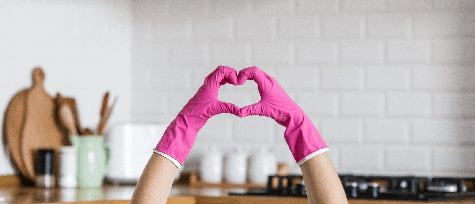Two hands making a heart whilst wearing hot pink kitchen gloves. Background shows a white kitchen wall with wooden countertop.