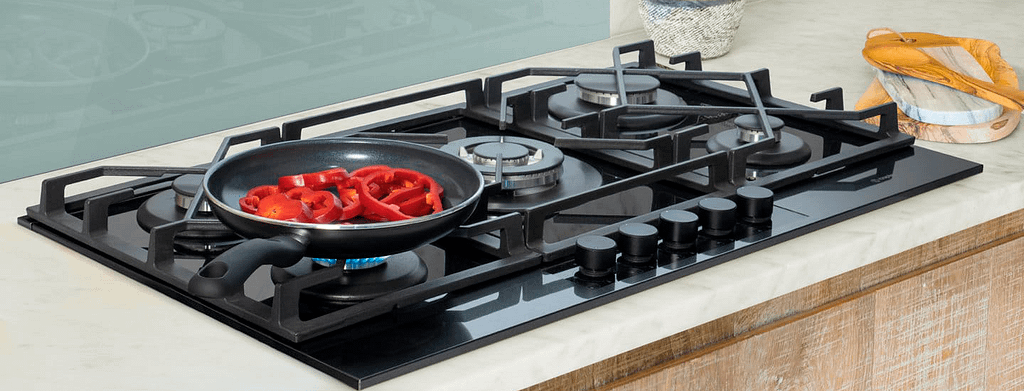 A gunmetal stainless steel hob with 5 burners, with a frying pan full of sliced red peppers.