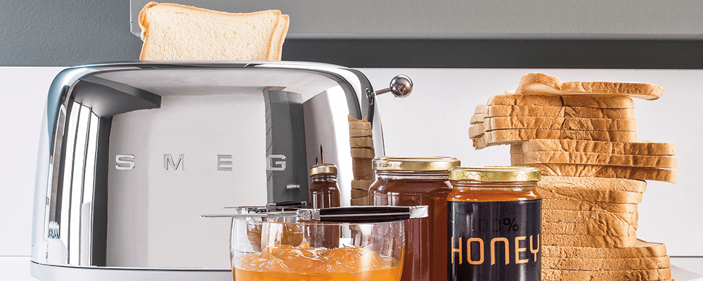 A chrome Smeg toaster with bread loaded, surrounded with bread, honey and marmalade.