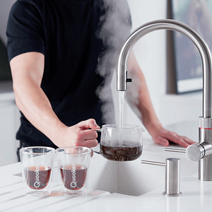 A Quooker boiling water tap in use. A man in a black T shirt is pouring boiling water into a teapot. Two hot coffees sit next to the sink.